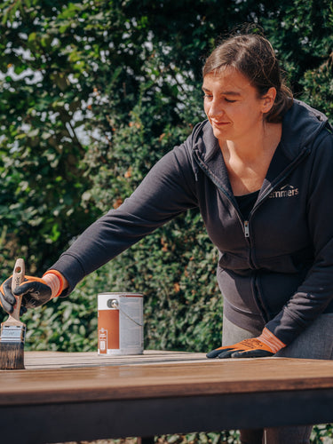 Eine Frau pflegt einen Gartentisch aus Holz mit einem Pinsel und schützendem Remmers Pflege-Öl.