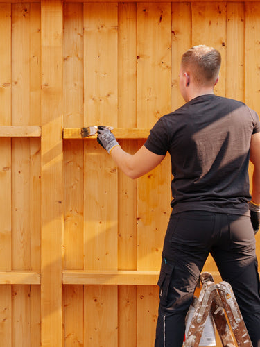 Eine Holzwand wird mit einem Pinsel und Remmers HK-Lasur für langlebigen Wetterschutz gestrichen.